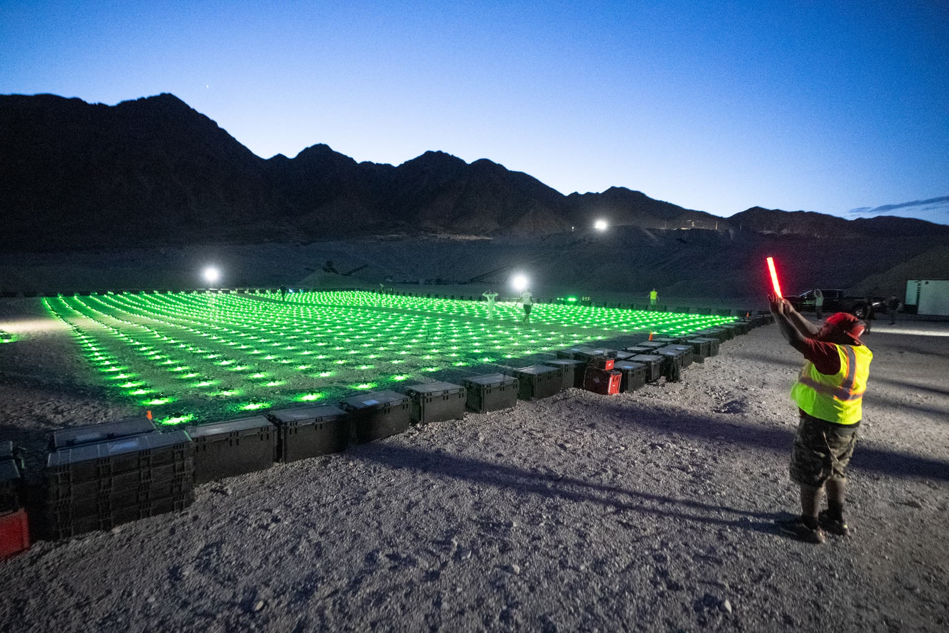 A large grid of green-lit drones with a crew member holding a light wand beside the launch area