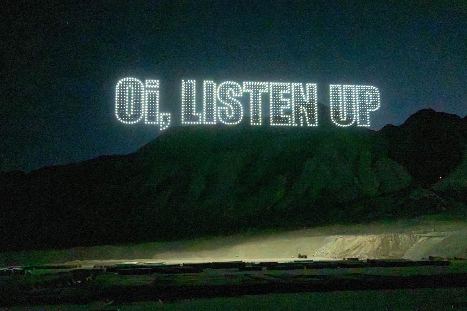 Drones spelling Oi, Listen Up in bright lettering above the mountains